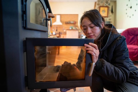 An Asian female looks down while meticulously cleaning the glass door of a wood burning stove in a