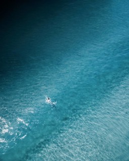 An aerial shot of a lone surfer paddling through calm turquoise waters along the Portuguese coast.