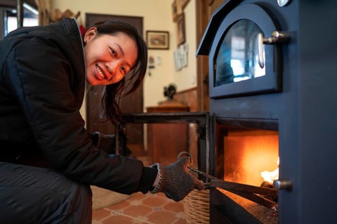 An Asian woman with visible joy looks directly at the camera while tending a wood burning stove