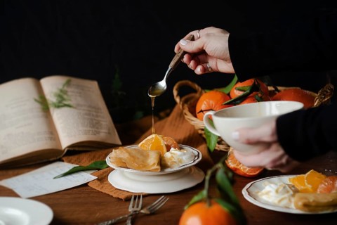 Cropped unrecognizable person drizzling honey over citric crepes, accompanied by fresh orange