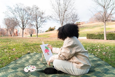 A young black woman sits on a blanket in a park, painting a colorful tree on a canvas. Surrounded