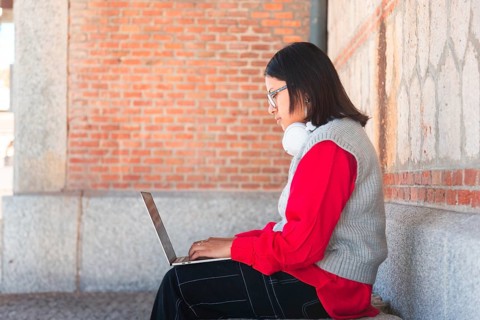 A Hispanic student with headphones focuses intently on her laptop while sitting against a brick