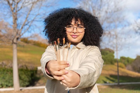 A joyful black woman with glasses and curly hair holds paintbrushes in a sunny park. The background