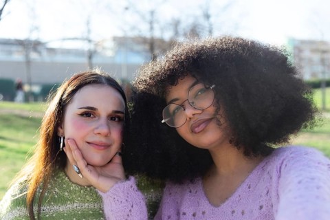 Two friends pose happily in a sunny park. One gently supports the other chin with a hand. They wear