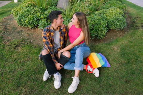 A joyful lesbian couple sits closely together on grass, embracing lovingly. A vibrant pride flag