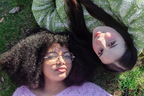 Two friends relax on grass, enjoying the sunshine. One wears glasses and has curly hair, the other