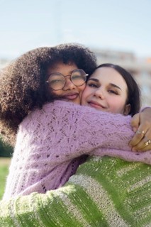 Two friends share a heartfelt hug, beaming with joy in a sunny outdoor setting. Both wear cozy