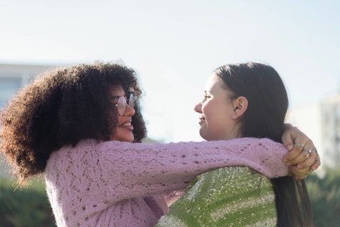 Two friends share a warm embrace in a sunlit park, wearing cozy sweaters. Smiling and enjoying each