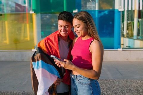 A happy lesbian couple looks at a smartphone together, smiling. One partner is wrapped in a Pride