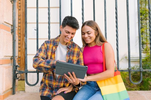 A happy lesbian couple sits together on a bench, smiling while looking at a tablet. One partner