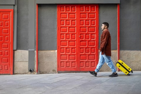 A latino transgender man walks confidently past vibrant red doors, pulling a bright yellow suitcase