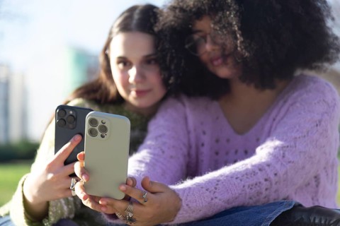 Two friends are sitting outdoors, capturing a selfie with smartphones. They are enjoying a sunny