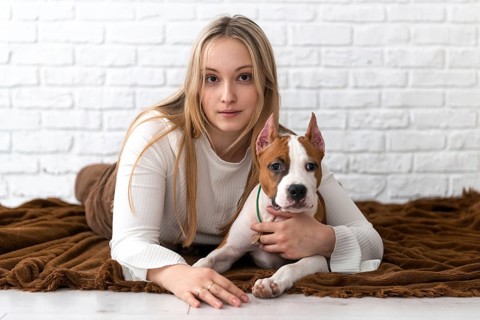 A young woman with blonde hair lounges on a brown blanket, affectionately cuddling her white and