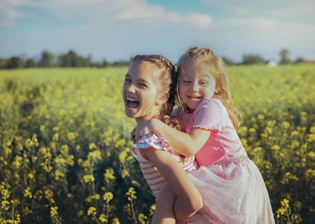 A young girl carries her little sister on her back as they laugh together in a field of bright