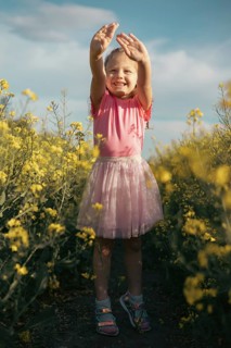 A joyful young girl in a pink outfit lifts her hands towards the sky while standing in a sunlit