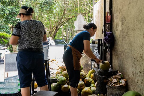Two women prepare coconuts at a street stall, using a machete to open them. Fresh coconuts pile in