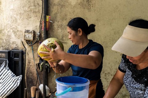 Two women skillfully open a coconut, letting its water drain into a blue bucket. The scene is set
