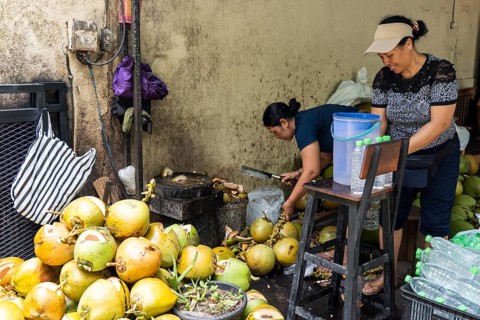 Two women processing coconuts in an urban setting. One uses a machete to expertly open coconuts,