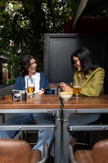 A lesbian couple enjoys drinks at an outdoor cafe. They smile and chat, surrounded by greenery,