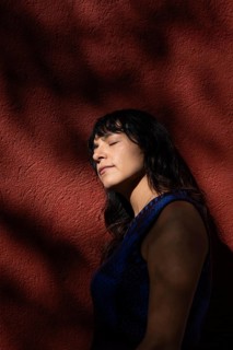 A Latin woman leans against a red wall with her eyes closed, basking in the sunlight. Shadows from