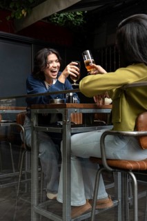 A lesbian couple joyfully toasts with glasses at a cozy cafe patio. They share a moment of laughter