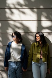 A lesbian couple stands against a wall, holding hands and enjoying the dappled sunlight. Both wear