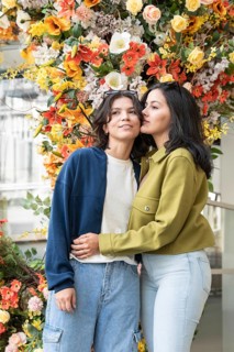 A lesbian couple shares a tender moment in front of a vibrant floral backdrop. Their closeness and