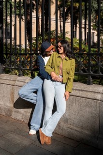 A joyful lesbian couple stands close, smiling and holding hands in a sunlit urban setting. They