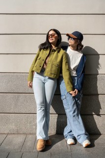 A lesbian couple stands outside, holding hands and enjoying the sunlight. Dressed in casual attire,
