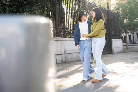 A lesbian couple walks hand in hand on a sunny urban street, embracing and smiling at each other.