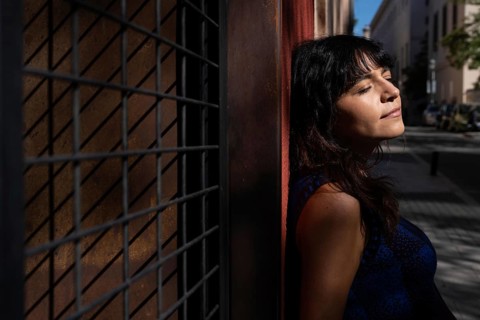 A Latin woman leans against a textured wall with her eyes closed, enjoying the warmth of the sun.