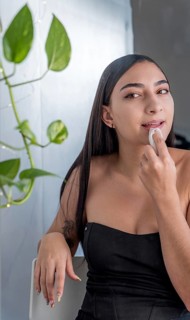 A woman is seated, applying makeup with a sponge in a softly lit room. She wearing a black top,