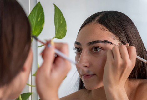 A woman applies makeup with a brush, concentrating on her reflection in a mirror. A green plant