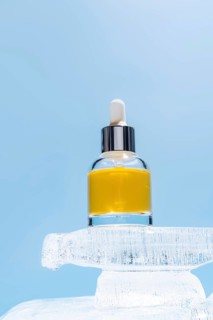 A cosmetic serum bottle with a dropper perched atop a block of ice, highlighted against a soft blue