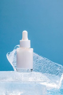 A single serum bottle with a dropper rests on clear ice chunks against a vibrant blue background,