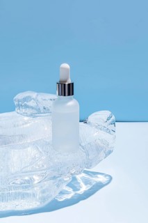 A frosted glass serum bottle with a dropper, resting in a carved ice block on a vibrant blue