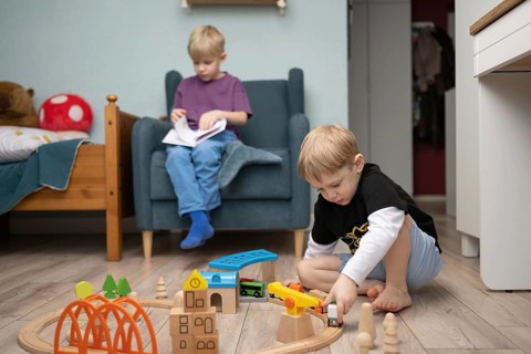 Two young children in a cozy room, one plays with wooden train toys on the floor, while the other