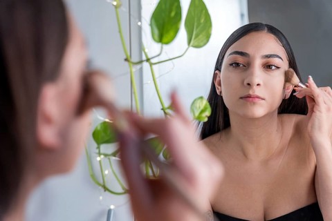 A woman carefully applies makeup using a brush in front of a mirror for the makeup session. Her