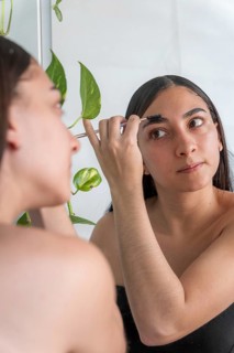 A woman focuses on applying makeup with a brush in front of a mirror for the makeup session. A
