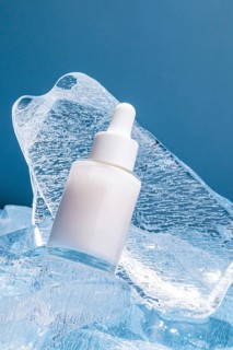 A serum bottle with a dropper rests on textured melting ice cubes against a blue background,