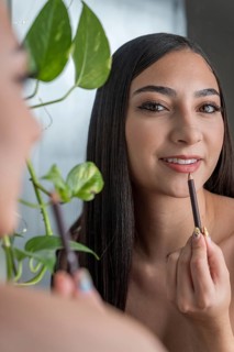 A woman applies lip liner while smiling at her reflection in a mirror in makeup session. A green