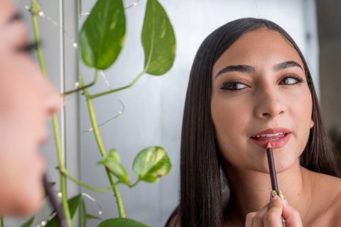 A woman applies lip liner while looking in a mirror, with lush green leaves in the background. The
