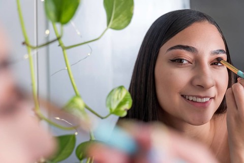 A woman is seen smiling while applying eye makeup in front of a mirror for the makeup session. A