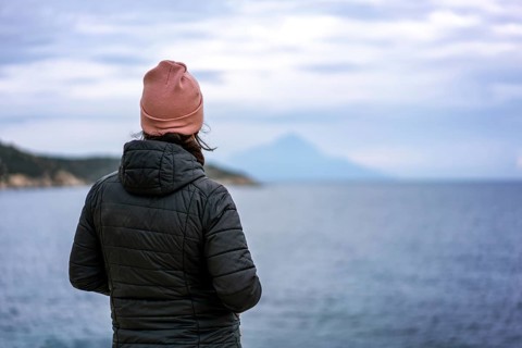 A person in a pink beanie and black jacket stands by the sea in Greece, gazing at the horizon. The