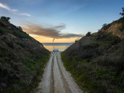 A narrow road flanked by green hills leads to a tranquil beach in Greece. A van is parked near the