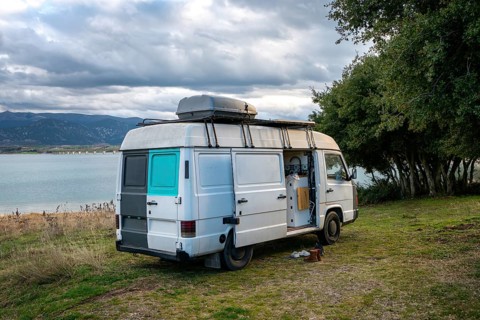 Camper van parked beside a scenic lake in Greece, surrounded by lush greenery and mountains. Cloudy