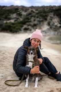 A woman wearing a pink beanie and glasses sits on a sandy beach in Greece, hugging a dog. The