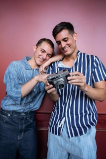 Charming gay couple smiling while reviewing photos on a vintage camera against a red wall. Casual