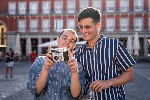 Happy gay couple reviewing photos on a vintage camera in Madrid's Plaza Mayor. Casual fashion,