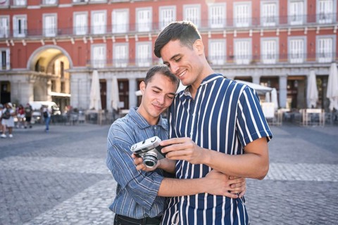 Gay couple embracing while reviewing photos on a vintage camera in Madrid's Plaza Mayor. Warm